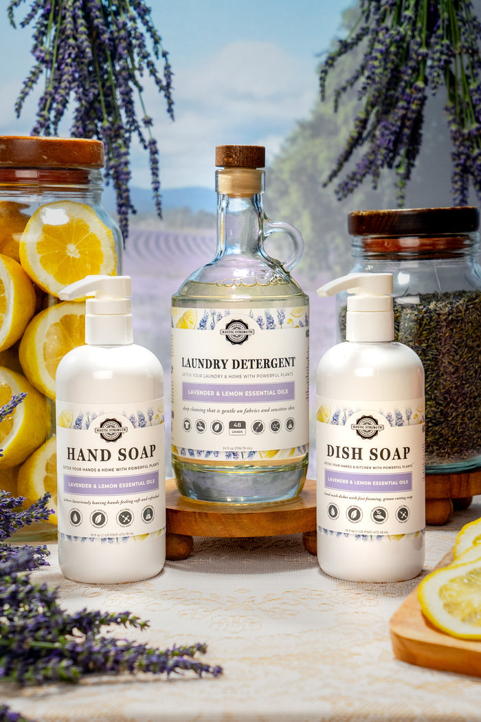 Rustic Strength - Cleaning Set | Lavender & Lemon containing a 24oz Glass | Laundry Detergent, 16oz Plastic |  Hand Soap, and a 16oz Plastic | Dish Soap. Lavender buds and lemon slices litter the background.