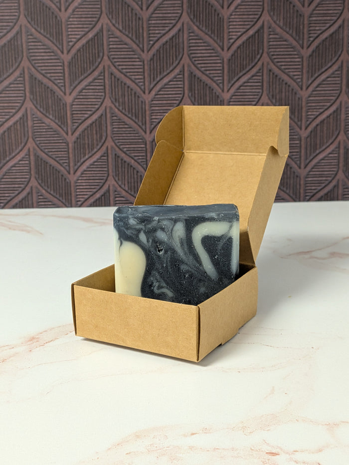Rustic Strength, Bar Soap, Charcoal Detox, a black & white swirled bar sits inside an open brown kraft paper box on a light marble surface, with a patterned dark wall in the background.