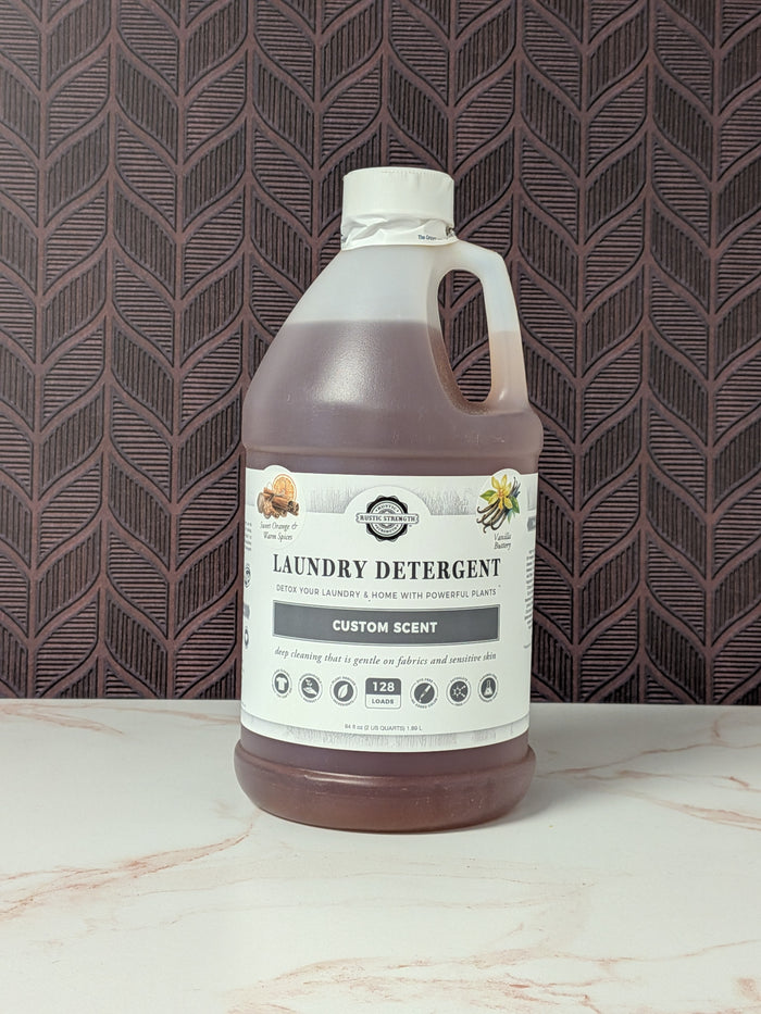 Rustic Strength, Laundry Detergent, Half Gallon, Plastic Jug, Sweet Orange & Warm Spices & Vanilla Buttery set on a light marbled surface with a patterned backdrop
