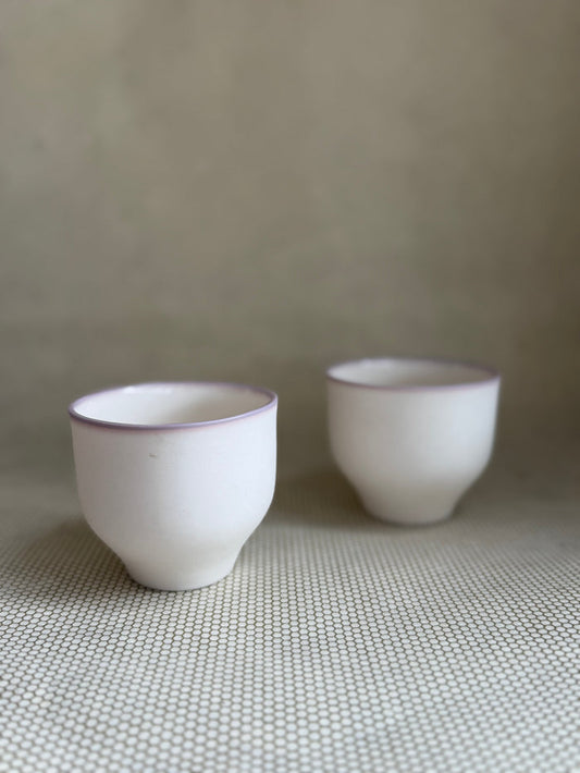 Two white porcelain cups with lavender rims on a surface with a neutral background.